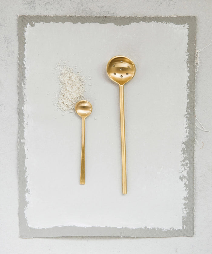 A minimalist brass slotted spoon with a long handle and round, perforated ladle styled with a small brass spoon, evoking elegance and simplicity. | Wildwood Home Co.